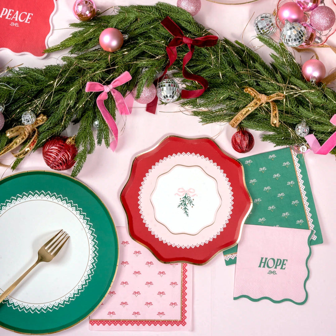 Christmas-themed table setting with plates, napkins, and ornaments on a pink background.